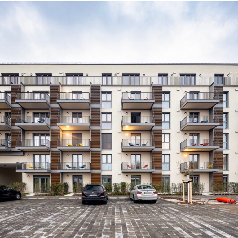 Außenansicht eines modernen Mehrfamilienhauses mit beiger Fassade und vielen Balkonen. Die Balkone haben schwarze Metallgeländer und sind mit vertikalen, braunen Holzelementen akzentuiert. Im Vordergrund ein Parkplatz mit dunklem Pflaster.