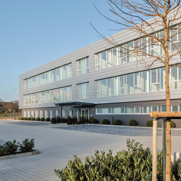 Modernes Büro- oder Geschäftsgebäude mit drei Stockwerken, einer grau-silbernen Fassade und horizontalen Fensterbändern. Das Gebäude steht auf einem großen gepflasterten Vorplatz mit niedrigen Büschen.