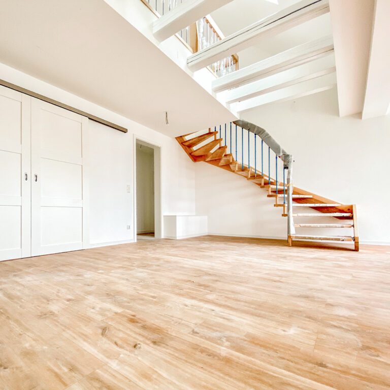 Innenansicht einer sanierten Wohnung mit hellem Holzboden, weiß gestrichenen Wänden und Holzbalken. Eine freistehende Holztreppe mit blauen Geländerstäben führt in eine obere Etage oder Galerie.