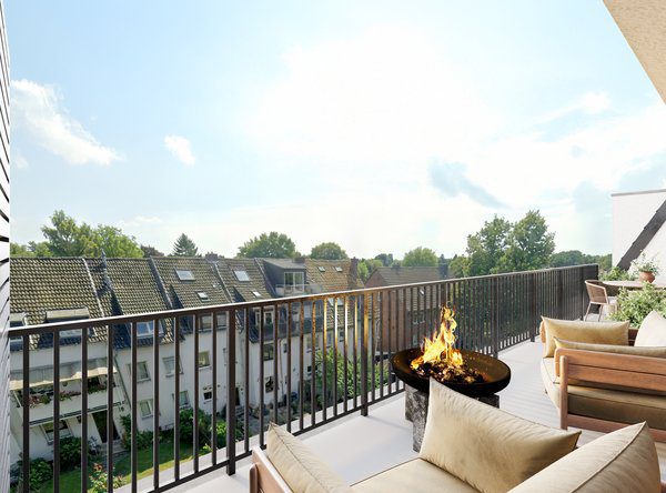 Ausblick von einer sonnigen Loggia oder Dachterrasse über ein dunkles Metallgeländer hinweg auf eine grüne Umgebung mit einer gegenüberliegenden Wohnhauszeile. Im Vordergrund brennt ein gemütliches Feuer in einer stilvollen, dunklen Feuerschale, umgeben von modernen Outdoor-Sesseln mit hellen, beigen Kissen.